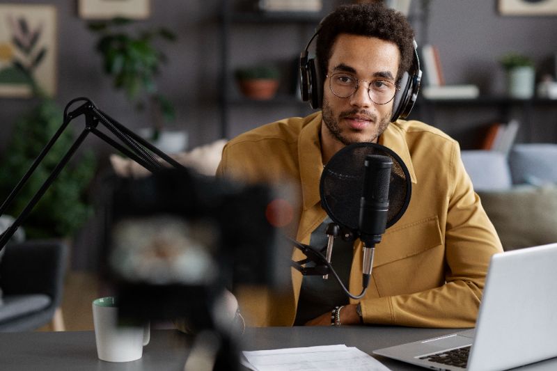 Estúdio de rádio moderno com mesa de som, fones de ouvido, microfones prateados e tela de computador com gráficos de áudio e ícones de podcasts.