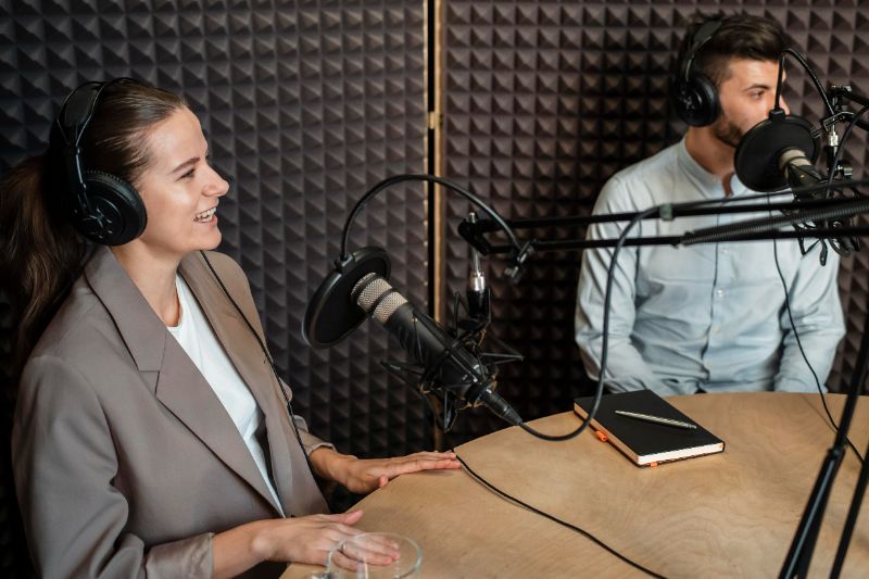 Microfone prateado em estúdio de rádio com fones de ouvido coloridos e tela com formas de onda no fundo.